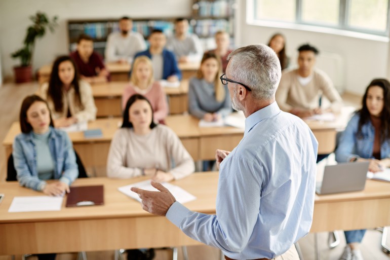 Back view of mature professor giving lecture to large group of college students in the classroom.; Shutterstock ID 2112409937; purchase_order: aljazeera ; job: ; client: ; other: