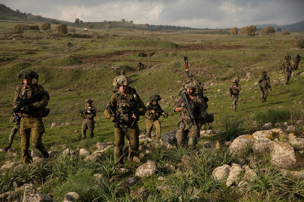 Soldiers walk across a grassy and rocky field carrying weapons.