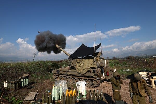 A plume of black smoke bursts forth as a tank fires artillery. A solder covers his ears and another turns away. In the foreground ammunition is set up.