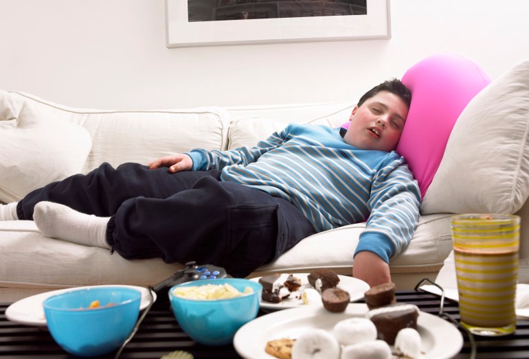 Young, Overweight Boy Sleeps on a Sofa Next to a Table of Crisps and Biscuits