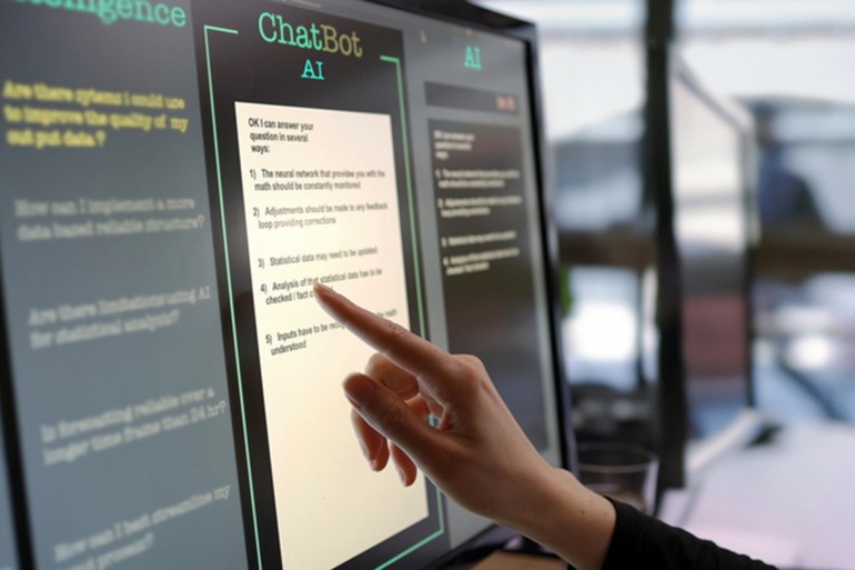 Close-up stock photograph showing a touchscreen monitor being used in an open plan office. A woman’s hand is asking an AI chatbot pre-typed questions & the Artificial Intelligence website is answering.