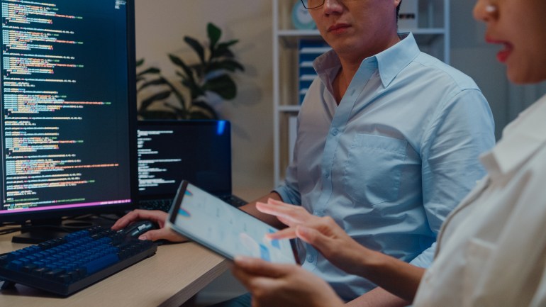 Closeup group of Asian people software developers using computer to write code sitting at desk with multiple screens in office at night. Programmer development concept.