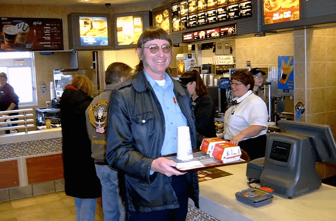 Donald Gorske holding a tray in McDonald's. He has straight-across bangs, long hair and is wearing a blue shirt with a leather jacket and sunglasses.