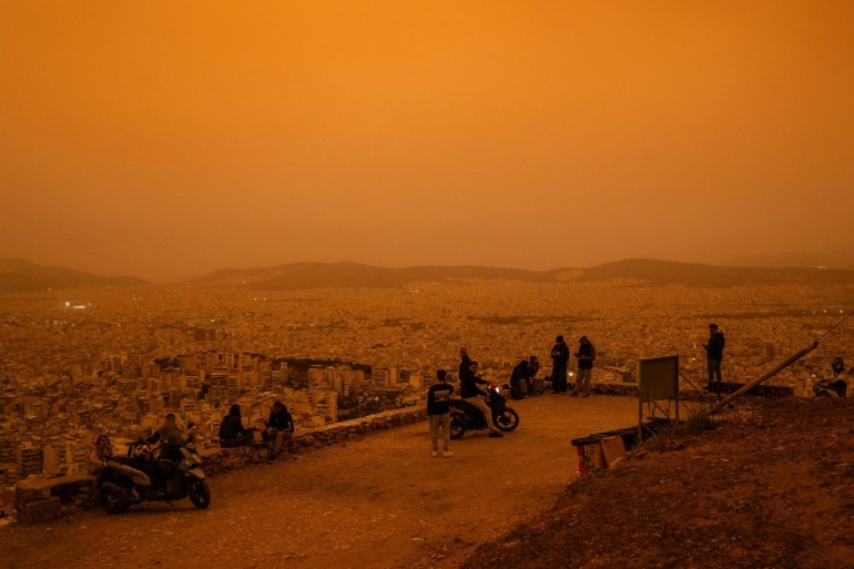 People sit on Tourkovounia hill overlooking the city of Athens, as southerly winds carry waves of Saharan dust to the city, in Athens, on April 23, 2024. - Clouds of dust blown in from the Sahara covered Athens and other Greek cities on April 23, 2024, one of the worst such episodes to hit the country since 2018, officials said. The yellow-orange haze smothered several regions, limiting visibility and prompting warnings of breathing risks from the authorities. (Photo by Angelos TZORTZINIS / AFP)