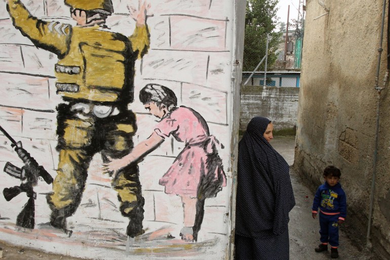 امرأة فلسطينية تقف مع طفل في زقاق بجوار نسخة عن جدارية «حارس الضفة الغربية» لبانكسي في مخيم العروب (الفرنسية)